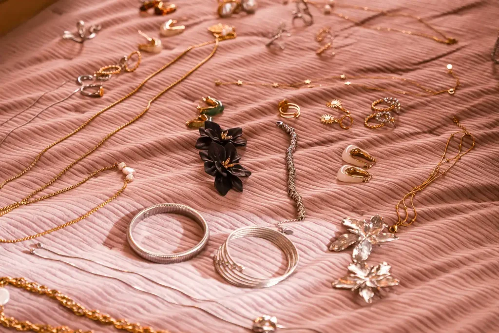 Composición flatlay sobre tela rosa mostrando la variedad de la colección Vousha: brazaletes plateados tipo cable, aretes de flores negras mate y collares de perlas.