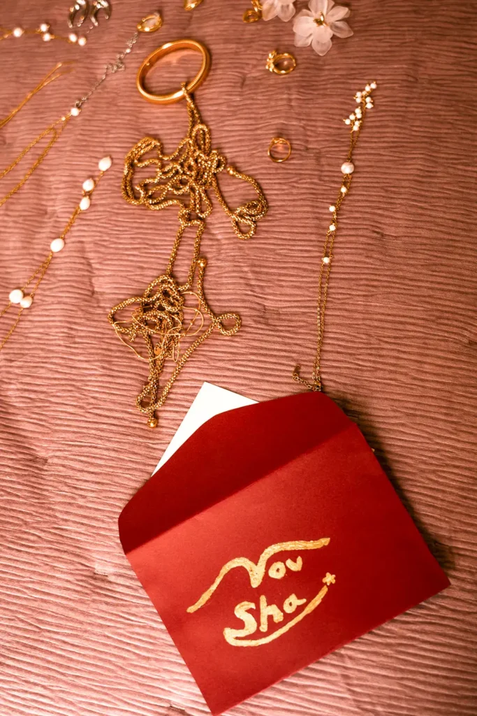 Composición de collares de perlas, brazaletes dorados y anillos sobre textura rosada junto a un sobre rojo con caligrafía dorada.